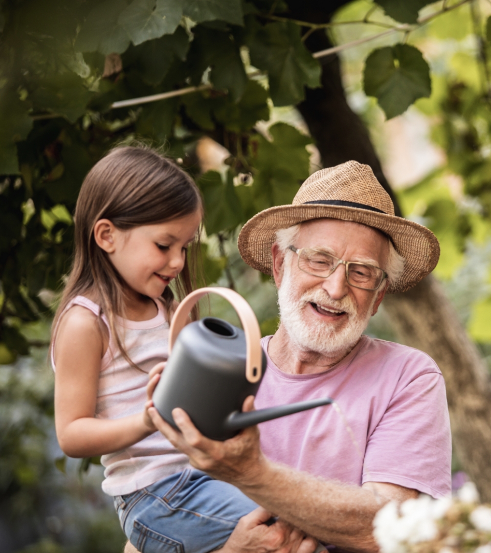 Photo of an older man carrying a young girl, helping her to water plants with a watering can.